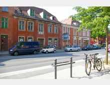 Straßenszene mit historischen Ziegelgebäuden im Vordergrund, darunter ein mehrstöckiges Haus mit grünen Fenstern und alten Fassaden. Mehrere geparkte Autos und Fahrräder befinden sich entlang der Straße, die von einem Bürgersteig und Bäumen gesäumt wird.