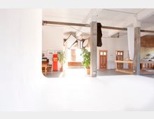 Großer, heller Raum mit industriellem Charakter und Betonpfeilern, der teilweise als Fotostudio genutzt wird. Im Hintergrund befinden sich Holztüren, Sofas, Topfpflanzen, ein roter Kühlschrank, Regale und Studiobeleuchtung; die Wände sind teilweise aus Ziegelstein.