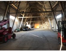 Große, leere Scheune mit Sicht auf ein offenes Holzgebälk und Steinwände. Im Innenraum stehen verschiedene Fahrzeuge, darunter ein roter Gabelstapler, sowie Werkzeug und Baumaterialien; im Hintergrund befinden sich große Holztore mit einfallendem Sonnenlicht.