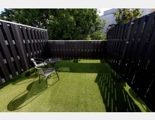 Eine kleine Terrasse mit Kunstrasenboden, umgeben von dunklen Holzzäunen. In der Mitte stehen ein runder, schwarzer Tisch und zwei dazu passende Metallstühle; im Hintergrund sind Bäume und teilweise Gebäude sichtbar.