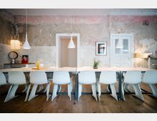 Langer Besprechungstisch mit weißen, modernen Stühlen in einem Raum mit rustikaler, unbehandelter Wandgestaltung. Hängelampen über dem Tisch, Holzboden und Türen mit klassischer weißer Zierverkleidung.