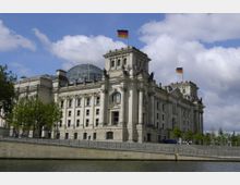 Das Bild zeigt das Reichstagsgebäude in Berlin, ein historisches Parlamentsgebäude mit prächtigen Fassaden, hoher Kuppel aus Glas und Stahl sowie gehissten deutschen Flaggen auf den Türmen. Im Vordergrund ist ein Fluss und eine Uferpromenade zu sehen.