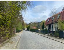 Pflasterstraße in einem Wohngebiet mit Einfamilienhäusern, einem Haus mit rotem Ziegeldach und Vorgarten auf der rechten Seite. Auf der linken Straßenseite stehen Bäume mit frischem Frühlingslaub und ein Zaun, im Hintergrund ist ein geparktes Fahrzeug zu erkennen.