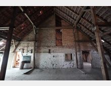 Innenraum einer alten Scheune mit hohen Holzbindern und sichtbarer Dachkonstruktion über einer teils weiß gestrichenen, teils unverputzten Backsteinwand. Mehrere Öffnungen und Türen führen in Nebenräume, der Boden besteht aus rohem Beton mit etwas Schmutz und Geröll.