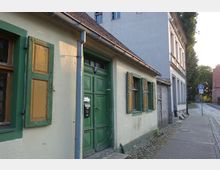 Kleines, historisches Gebäude mit gelber Fassade, grün gestrichenen Fensterläden und einer großen grünen Tür, entlang einer gepflasterten Straße; daneben ein größeres Haus in schlichter grauer Putzarchitektur. Ein Schild weist auf eine Parkzone hin, und im Hintergrund sind Bäume und weitere Gebäude sichtbar.