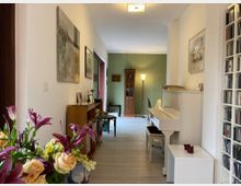 Wohnzimmer mit hellem Holzboden, weißen Wänden und einem grünen Akzentbereich. Zu sehen sind ein weißes Klavier, Bücherregale, Blumenarrangements auf einem Tisch und eine hohe Vitrine im Hintergrund.