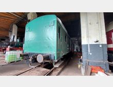 Grüner Zugwaggon in einer Lokhalle mit sichtbaren Schienen darunter; im Hintergrund stehen weitere historische Lokomotiven. Die Halle hat eine Holzdecke und gemauerte Wände, teils weiß gestrichen.