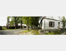 Straßenansicht in einem Dorf mit einer historischen Kirche aus Backstein auf der linken Seite, umgeben von einer Mauer, und einem weiß gestrichenen, alten Gebäude mit verwitterten Fenstern und Treppenstufen rechts. Zwischen Kirche und Gebäude verläuft eine asphaltierte Straße, gesäumt von hohen Bäumen.