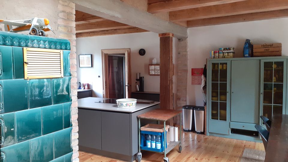 Rustikale Wohnküche mit sichtbaren Holzbalken, Holzfußboden und gemauerten Stützen. Links ein grüner Kachelofen, in der Mitte eine Kücheninsel mit Kochfeld und rechts ein großer Vitrinenschrank sowie ein Esstischbereich.