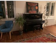 Ein Wohnzimmer mit Holzboden, einem schwarzen Klavier, auf dem ein Gemälde steht, einer Zimmerpflanze in einem Topf, einem blauen Sessel und einer Stehlampe mit runden Glaselementen. Zwei Fenster mit weißen Rahmen befinden sich im Hintergrund.