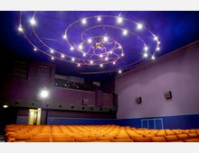 Kino-Innenraum mit orangefarbenen Sitzreihen und lila Wänden. Die Decke ist in Dunkelblau gestaltet und mit einer spiralförmigen Installation aus Lichtern dekoriert.