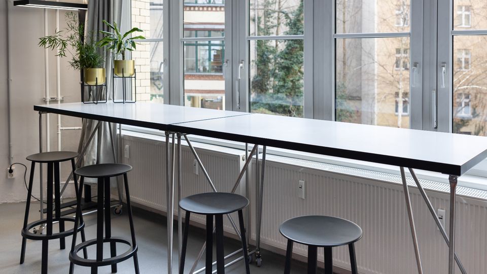 Heller Innenraum mit langem, schmalem Stehtisch auf Metallgestell und mehreren schwarzen Hockern entlang einer Fensterfront. Große, mehrteilige Fenster mit Blick auf benachbarte Gebäude; auf der Tischplatte stehen Pflanzen in Töpfen.