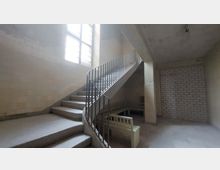 Treppenhaus in einem Gebäude mit kahlen Wänden und einer großen Fensterfront, die natürliches Licht hereinlässt. Eine schlichte Wendeltreppe mit Metallgeländer führt nach oben, daneben stehen helle Sitzbänke auf einem Steinboden.