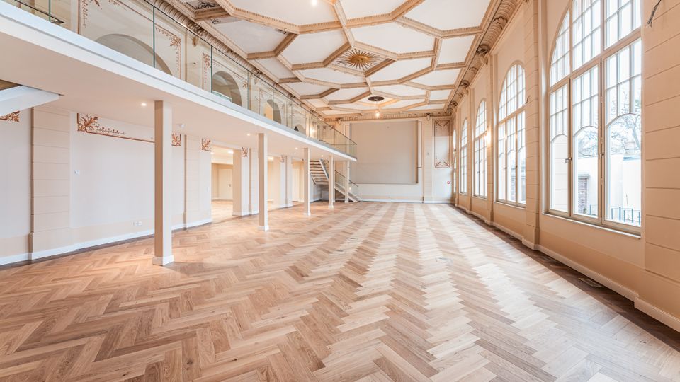 Großer, leerer Saal mit hellem Fischgrätparkett, hoher kassettierter Stuckdecke und umlaufender Empore mit Glasgeländer. Rechts sorgen hohe, bogenförmige Fensterfronten für viel Tageslicht; im Hintergrund führt eine Treppe zur oberen Ebene.