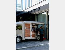 Ein silberner Foodtruck mit Holzverkleidung steht auf einem gepflasterten Gehweg vor einem modernen Gebäude mit Glasfassade. Eine Person bestellt an der Theke, auf der eine Menütafel mit handgeschriebener Schrift angebracht ist.