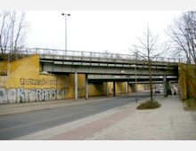 Straßenunterführung mit einer gelben Backsteinmauer, die mit Graffiti besprüht ist, und einer darüber verlaufenden Stahlkonstruktion aus Brückenelementen. Auf der Straße darunter sowie dem angrenzenden Gehweg sind wenige Menschen und Autos zu sehen, umgeben von kahlen Bäumen.