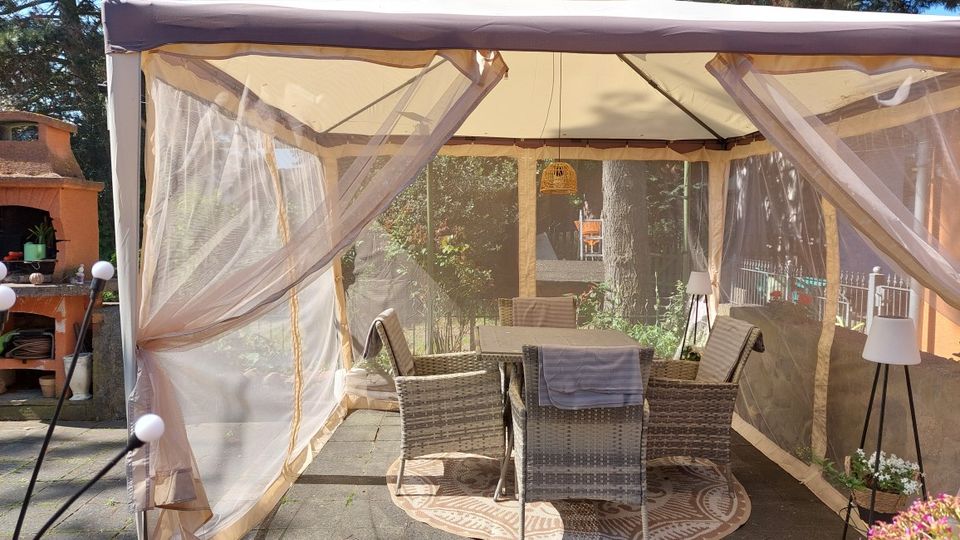 Überdachter Gartenpavillon mit beigefarbenem Stoffdach und transparenten Seitenwänden, innen ein Tisch mit mehreren Rattan-Stühlen auf einem runden Teppich. Links steht ein gemauerter Grillkamin auf der Terrasse, im Hintergrund sind Bäume und Gartenbewuchs zu sehen.