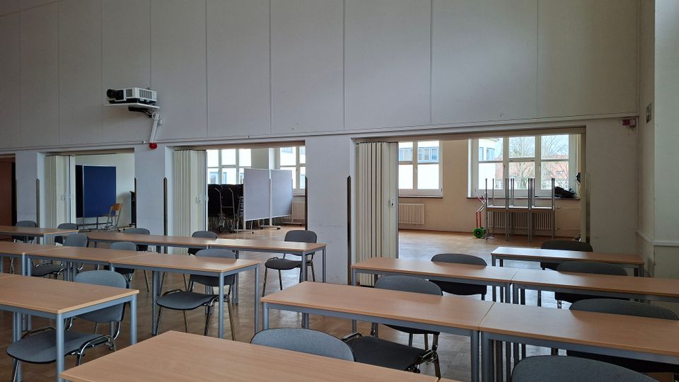 Großer Seminar- oder Klassenraum mit Reihen von rechteckigen Tischen und grauen Stühlen auf Parkettboden. Hinten befinden sich breite Durchgänge zu Nebenräumen sowie große Fenster mit Heizkörpern darunter und ein an der Wand montierter Deckenprojektor.
