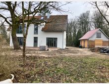 Ein weißes zweistöckiges Haus mit Satteldach und großen Fenstern steht in einer ländlichen Umgebung. Rechts daneben befindet sich ein kleineres Gebäude mit teils verklinkerten Wänden, noch im Bau oder in Renovierung, umgeben von kahlem Boden und Bäumen.