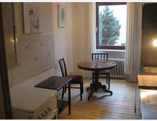 Kleine Küche mit Holzdielenboden, runden Holztisch und zwei Stühlen vor einem Fenster mit Blick auf einen Baum. Links ist eine geflieste Wand und ein Herd, rechts eine Arbeitsfläche und Spüle.