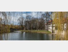 Flusslandschaft mit einer steinernen Bogenbrücke im Hintergrund, umgeben von kahlen Bäumen und angrenzenden Gebäuden mit roten Dächern und weißen Fassaden. Im Vordergrund hängen Zweige einer Trauerweide über das Wasser.