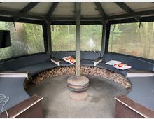 Rundes, überdachtes Gartenhäuschen mit umlaufender Sitzbank, dekoriert mit Kissen. In der Mitte ein kleiner Ofen, im Hintergrund außen Bäume und Büsche sichtbar.