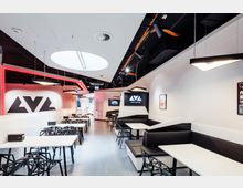 Ein moderner Gaming- und Lounge-Bereich mit weißen Tischen, schwarzen Stühlen und gepolsterten Eckbänken, beleuchtet durch Hängelampen und ein rundes Oberlicht. Rote LED-Leisten und Bildschirme mit dem Schriftzug "LVL world of gaming" ergänzen das futuristische Design.