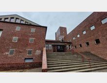 Backsteinbau mit modernistischem Design, bestehend aus mehreren Gebäudeteilen mit schlichten Fenstern. Eine breite Treppe mit rotem Geländer führt zum Eingangsbereich mit abgerundeter Überdachung, im Hintergrund ragt ein Turm empor.