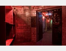 Industrieähnlicher Innenraum mit Betonwänden, graffiti-bedeckten Türen und rotem sowie blauem Licht, das eine düstere Atmosphäre schafft. Der Raum zeigt Schilder von Abnutzung, mit sichtbaren Leitungen an der Decke und einem Regal an der linken Wand.