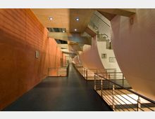 Breiter, moderner Innenraum mit langen, geraden Gehwegen, flankiert von rot-orangefarbenen Betonwänden auf der linken Seite und geschwungenen weißen Strukturen auf der rechten Seite, durchzogen von Metallgeländern und beleuchtet von eingebauten Deckenleuchten. Im Hintergrund sind Betonträger und Treppen sichtbar.