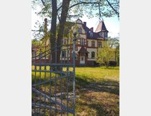 Historische Villa mit gelb-roter Fassade und spitzem Turmdach, umgeben von Bäumen und einer Rasenfläche, im Vordergrund ein geöffnetes schmiedeeisernes Tor.