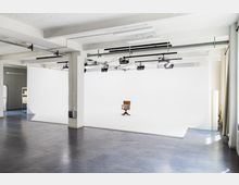 Ein geräumiges Fotostudio mit weißen Wänden und einem nahtlosen weißen Hintergrund, auf dem ein einzelner hölzerner Stuhl steht; an der Decke befinden sich mehrere Beleuchtungseinrichtungen. Der Boden ist mit einem dunklen, glatten Material ausgelegt, und das Studio hat eine offene, minimalistische Gestaltung.