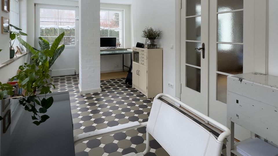 Heller, schmaler Innenraum mit gemustertem Fliesenboden in Grau- und Schwarztönen, weißer Decke und einem zentralen, weiß verkleideten Pfeiler. Am Ende stehen Schreibtisch und Schrank vor großen Fenstern; rechts sind doppelte Glastüren, links eine Fensterbank mit Pflanzen.