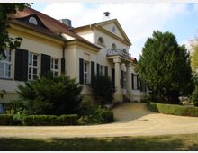 Ehemalige Villa in klassizistischem Stil mit cremefarbenen Wänden, schwarzen Fensterläden und einem roten Walmdach. Vor dem Gebäude führt ein geschwungener Weg durch einen gepflegten Garten mit Hecken und Bäumen.