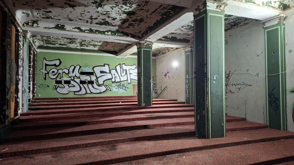 Innenraum eines verlassenen Gebäudes mit terrassenförmig ansteigendem Boden, mehreren grünen Stützpfeilern und einer stark abblätternden Decke. Wände mit Graffiti und verwitterter Farbe, seitlich eine Reihe hoher Fenster und einfache Türen.