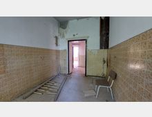 Ein verlassener, heruntergekommener Raum mit beigen Fliesen an den Wänden und einer beschädigten, weißen Decke. Im Raum stehen ein einzelner Stuhl und lose Holzstücke auf dem Boden, während durch eine offene Tür ein weiterer, heller Raum sichtbar wird.