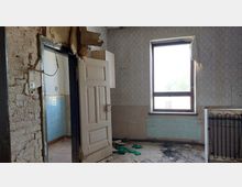 Ein verlassenes Zimmer mit sichtbaren Schäden: bröckelnde Tapeten, ein offenes Fenster mit Tageslicht, eine Heizkörperverkleidung und eine offene Tür, die in einen Raum mit blauen Wandfliesen führt. Die Wände sind teils unverputzt mit sichtbarem Mauerwerk, der Boden ist stark verschmutzt.