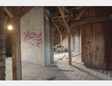 Dachboden eines alten Gebäudes mit Holzbalken, Holzfußboden und unverputzten Ziegelwänden. An einer Betonwand ist lila Graffiti, der Raum wirkt unfertig und teilweise verschmutzt.