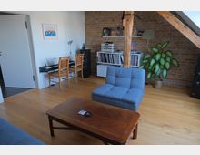 Wohnraum mit sichtbarer Backsteinwand, Holzbalken und Holzboden. Im Raum befinden sich ein Klavier mit zwei Stühlen, eine Musikanlage mit Lautsprechern und einem Regal voller Platten, eine Zimmerpflanze, ein blauer Sessel und ein Holztisch.