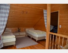 Schlafzimmer in einem Dachgeschoss mit Holzverkleidung an Wänden und Decke, ausgestattet mit zwei Einzelbetten, einem schlichten Teppich und Vorhängen an den Seiten. Im Vordergrund ist ein Holzgeländer zu sehen, und ein angrenzender Raum mit einer geöffneten Tür ist erkennbar.