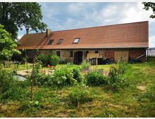 Backsteingebäude mit rotem Satteldach in einer ländlichen Umgebung, umgeben von einer Grasfläche und üppiger Vegetation. Im Vordergrund ein kleiner Gartenbereich mit Pflanzen und ein einfacher Teich.