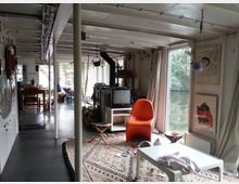 Ein Wohnbereich in einem Hausboot mit großen Fenstern, die auf das Wasser blicken. Der Raum ist mit modernen Möbeln wie einem orangefarbenen Stuhl, einem Holzofen, Teppichen und einem weißen Tisch ausgestattet; im Hintergrund sind eine Küchenecke und Esstisch sichtbar.