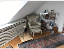 Wohnzimmer-Ecke unter einer Dachschräge mit einem floralen Sessel im Vintage-Stil, einem niedrigen Glastisch, einem Heizkörper mit weißer Abdeckung, einem schmalen Wandregal mit Dekorationen und Büchern sowie einem Teppich mit orientalischem Muster auf einem Holzboden.