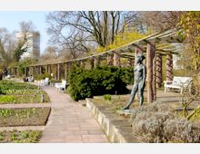 Ein Gartenweg mit Pflastersteinen führt an Blumenbeeten, weißen Bänken und einer Pergola mit steinernen Säulen vorbei. Im Vordergrund steht eine bronzene Skulptur vor Sträuchern und karger Vegetation, während im Hintergrund ein Hochhaus zwischen Bäumen sichtbar ist.