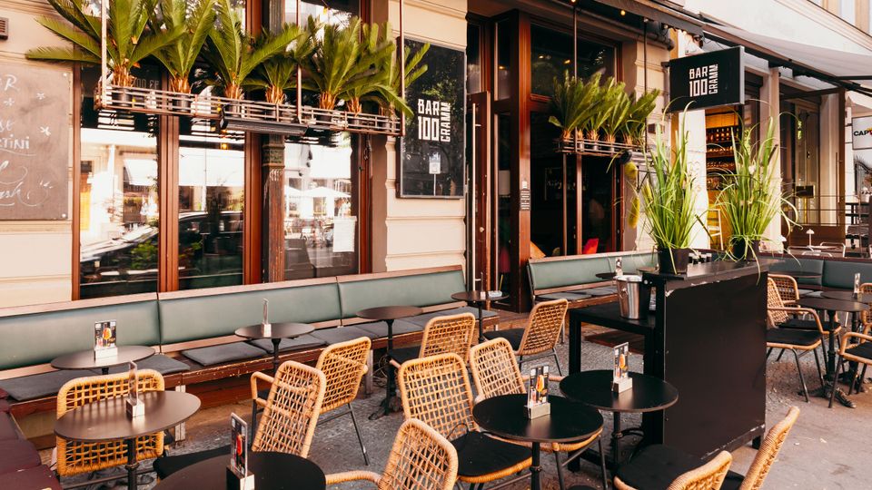 Außenbereich eines Straßencafés mit mehreren runden Tischen und Flechtstühlen sowie einer langen gepolsterten Sitzbank entlang der Fassade. Über den Fenstern hängen Pflanzkästen mit Palmen, und Markisen sowie ein Schild „Bar 1000“ rahmen den Eingangsbereich.
