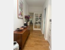 Ein schmaler Flur mit hellem Holzboden und weißen Wänden, auf der linken Seite ein Holzsideboard mit Dekorationen wie Blumenvasen und Schalen, darüber ein Bild an der Wand. Am Ende des Flurs eine verglaste Schiebetür, durch die ein weiterer Raum mit Regalen sichtbar ist.