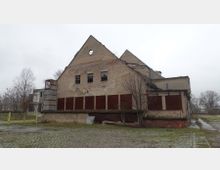 Zweistöckiges, verfallenes Gebäude aus Ziegelmauerwerk mit teils eingestürztem Dach, zugemauerten Fenstern und einer metallischen Anbaukonstruktion. Im Vordergrund ein gepflasterter Hof mit Grasbewuchs und vereinzelten kahlen Bäumen, umgeben von einer tristen Landschaft bei bedecktem Himmel.