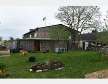 Ein großer Garten mit Rasenfläche, Blumenbeeten, Hochbeeten und jungen Bäumen vor einem älteren Gebäude mit einer Veranda. Oben weht eine ukrainische Flagge, und im Garten stehen Holzstühle, Gartenwerkzeuge und verschiedene Pflanzenbehälter.