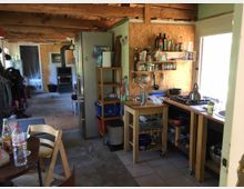 Rustikale Küche und Wohnbereich in einem Holzhaus mit sichtbaren Holzbalken an der Decke. Die Küche umfasst offene Regale, einen Herd mit Arbeitsfläche und einen kleinen Tisch mit Stühlen, während der Wohnbereich im Hintergrund einen Holzofen zeigt.