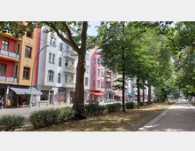 Straßenszene mit mehrfarbigen Altbauten, darunter Läden und Cafés mit Markisen, auf der linken Seite und einer parkähnlichen Grünfläche mit Bäumen und einem Gehweg auf der rechten Seite. Menschen gehen oder sitzen in der Umgebung, während sich Autos auf der Straße bewegen.